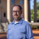 Nathan Lewis, a man with glasses and a beard wearing a light blue shirt, stands outside in front of a building with columns.