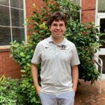 A person wearing glasses and a white Georgia polo shirt stands outside near green bushes and a brick building, smiling at the camera.