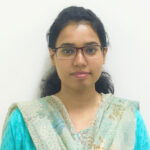 A woman with glasses and dark hair, wearing a light green printed dupatta over a matching kurta, poses in front of a plain white background.