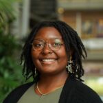 A person with glasses and shoulder-length locs smiles at the camera, wearing a green top, black cardigan, and decorative earrings. Indoor setting with blurred background.
