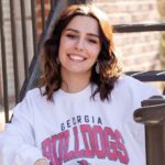 A young woman with shoulder-length brown hair smiles while sitting on outdoor stairs, wearing a white Georgia Bulldogs sweatshirt.