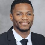 A man in a dark suit, white shirt, and black tie smiles slightly while posing for a professional headshot against a gray background.