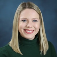 A woman with straight blonde hair, wearing a green turtleneck sweater, smiles in front of a blue studio background.