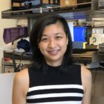 Kimberly Tsui, a woman with shoulder-length black hair and a sleeveless black and white striped top, stands smiling in an office or lab with shelves and assorted items behind her.