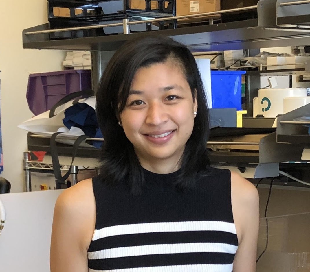 Kimberly Tsui, a woman with shoulder-length black hair and a sleeveless black and white striped top, stands smiling in an office or lab with shelves and assorted items behind her.