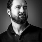 Black and white portrait of a bearded man with short, dark hair, wearing a buttoned sweater, looking slightly to the side against a plain background.