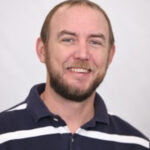 A man with short hair and a beard is smiling, wearing a navy blue polo shirt with white horizontal stripes against a plain light background.
