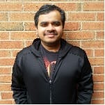 A person with short dark hair and a trimmed beard is wearing a black jacket and standing in front of a red brick wall, smiling at the camera.